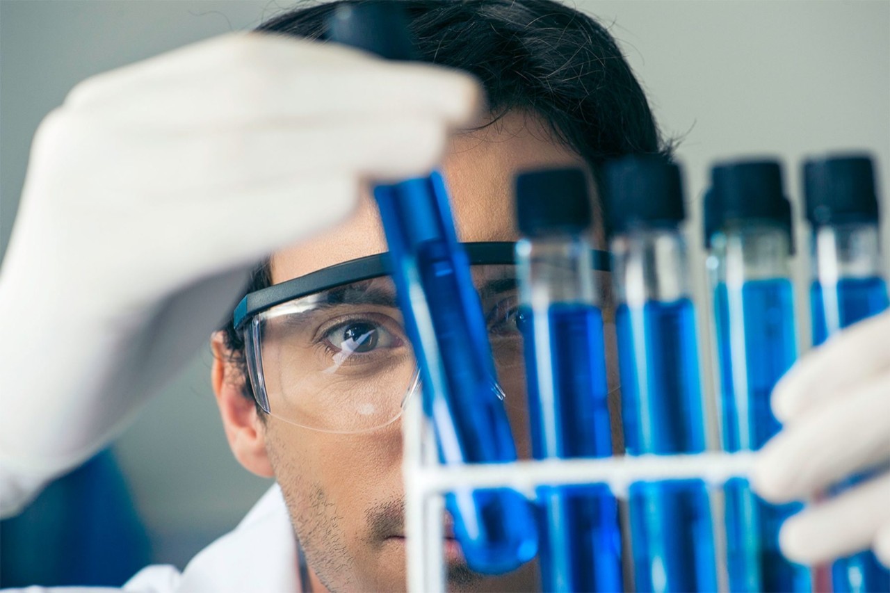A man in gloves and goggles looking closely at several test tubes containing blue liquid.